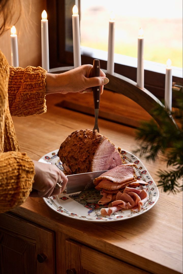 A woman slices a traditional Nordic Christmas ham in a window next to a Christmas candle arch. 