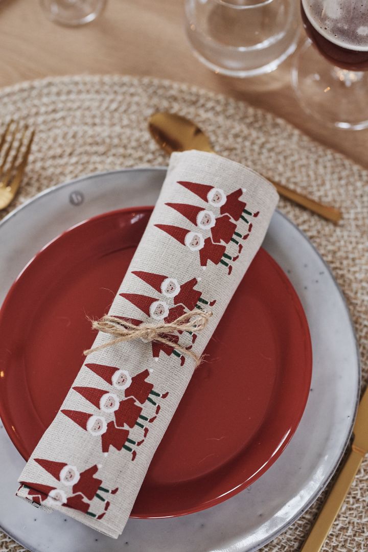 A rolled up tea towel with Santa Claus motifs rests on a red plate and a Nordic sand plate from Broste Copenhagen.