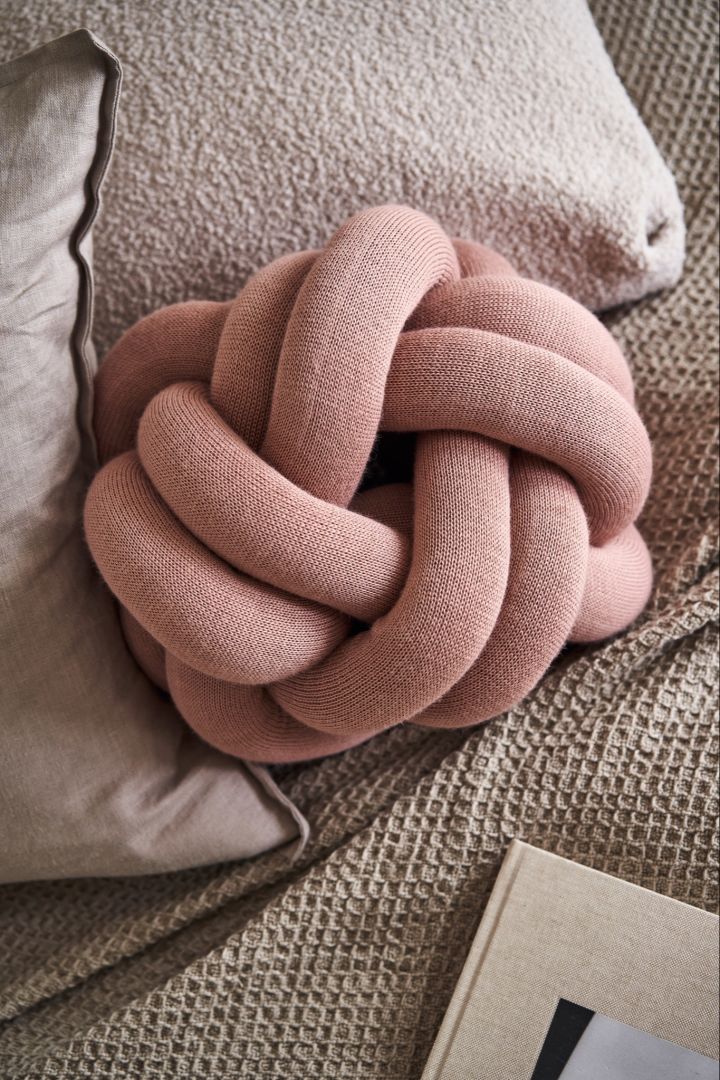 The Knot Cushion small in dusty pink sitting on a bed with beige and grey bed sheets. 